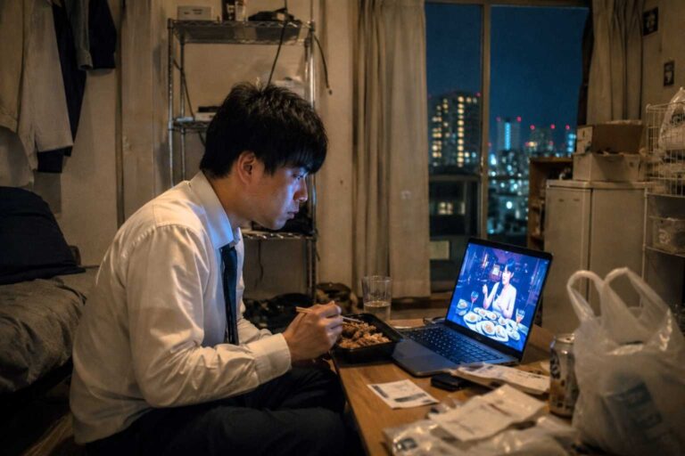 A man eats an expensive dinner alone in a small apartment while watching a food livestream.