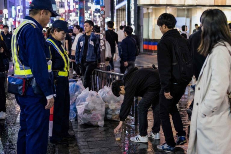 City patrol staff monitor a crowded Shibuya sidewalk as pedestrians pass with drinks and trash.